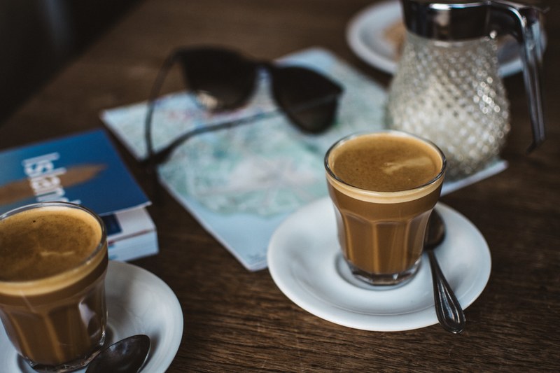 latte in a clear glass on a white saucer with a spoon with sunglasses and a map on the table, first coffee after arriving to iceland 4k