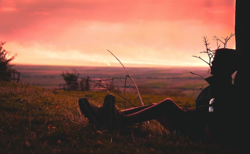 silhouette of a person sitting against a tree on a grassy field an orange and yellow sunset in the background, sunset tree silhouette and alone 4k