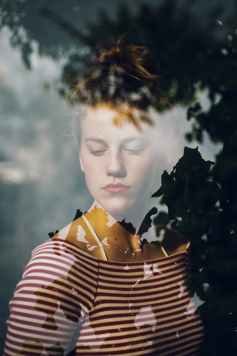 a person in an off the shoulder shirt looks down behind a window which reflects trees, double exposure 4k