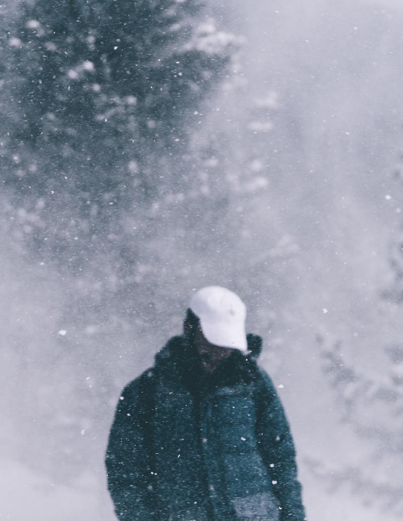 a person wearing a white hat and winter jacket looking down while walking through a forest amidst snowfall, walking in forest snowfall 4k