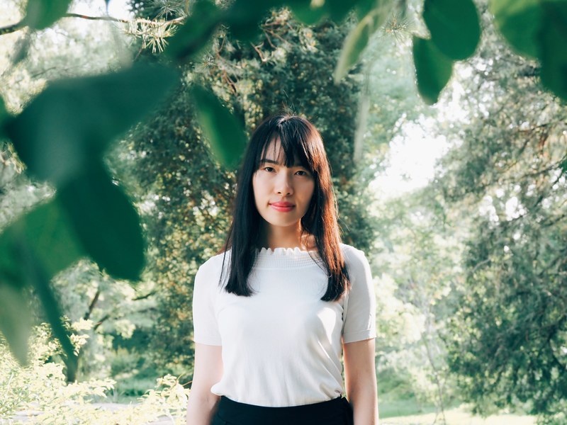 a smiling woman in a white shirt stands beneath sunny green trees, woman beneath trees and sun 4k