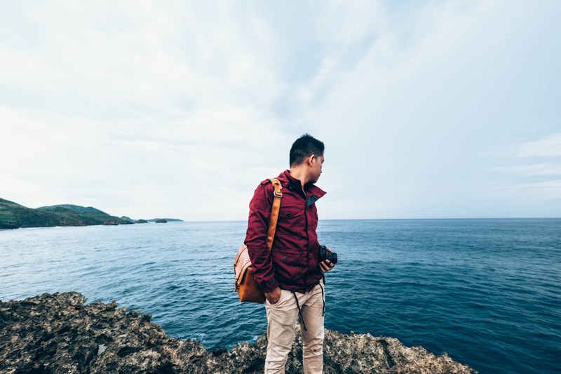 a man wearing a red jacket holds a camera and bag by the coast, man by the coast 4k