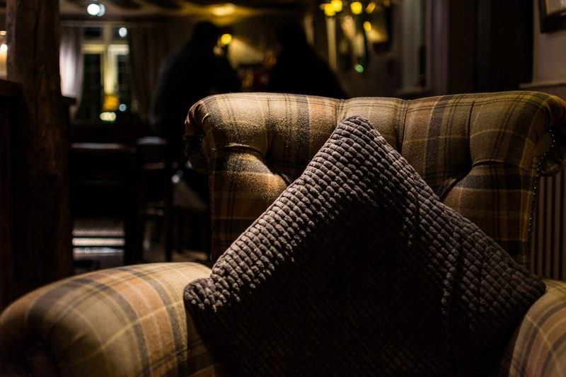 tartan chair with a gray pillow in a dim room with two people sitting in the background, pillow on an armchair 4k