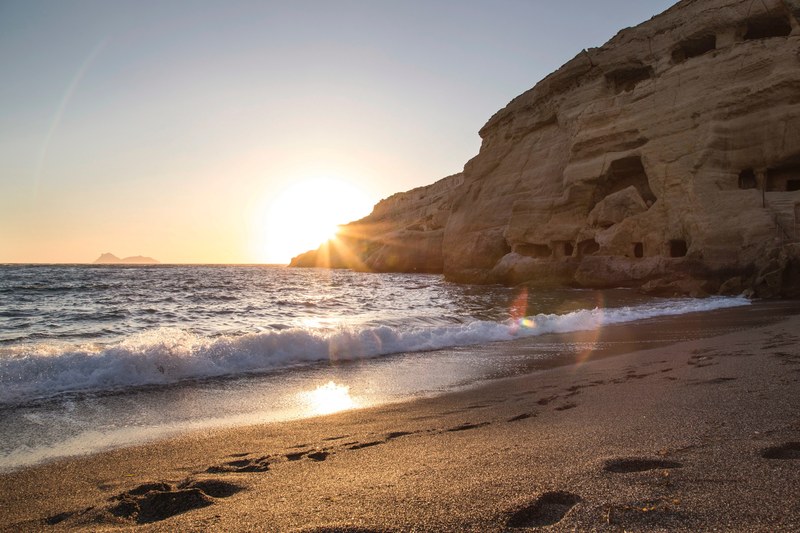 a course sand beach in matala with foot marks along the shoreline leading toward a large rock coastline_ small waves crash into shore and the bright sunrise or sunset creates a yellow glare 4k