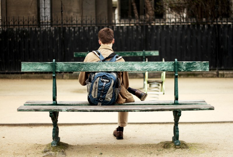 a person in a backpack sits on an outdoor bench crossing their legs, person sitting on a bench 4k