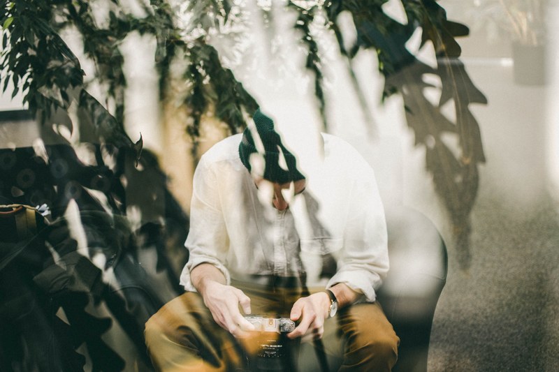 a man sits looking down at a camera behind a window which reflects trees outside, man with camera behind window 4k