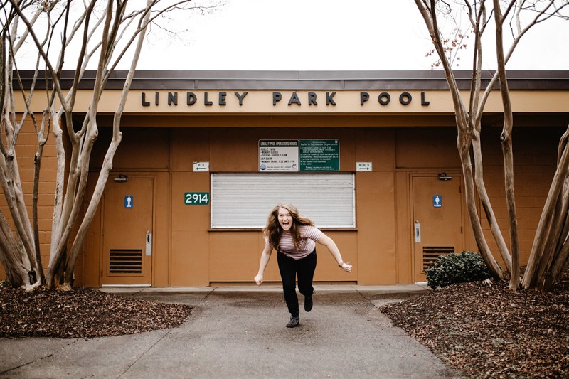 a woman laughs while lunging forward in front of the bathrooms at the lindley park pool, be happy 4k