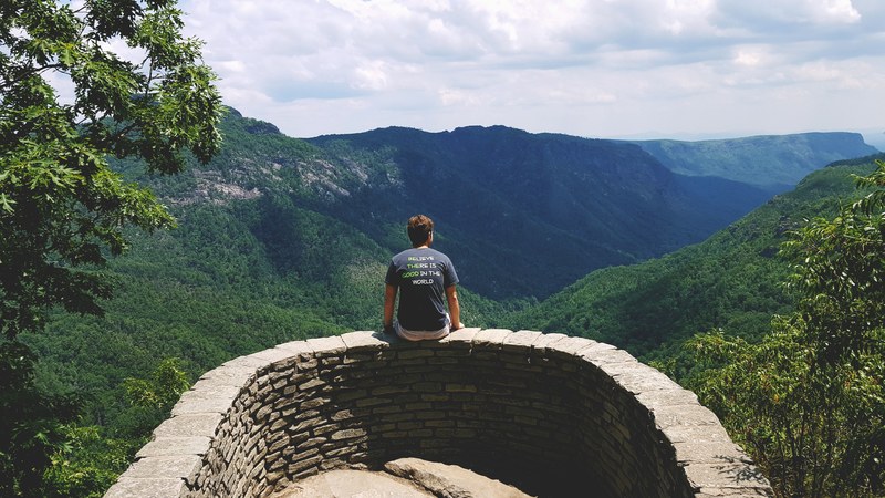 a man sitting on a small stone wall overlooking a green mountain valley, small wall by a mountain valley 4k