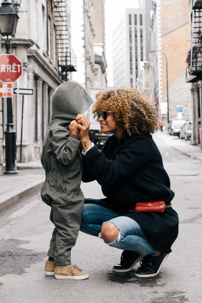 a mom with curly hair crouching in the street old port of montreal smiling at her young child wearing an all in one outfit with the hood up, smiles in the street from mom 4k