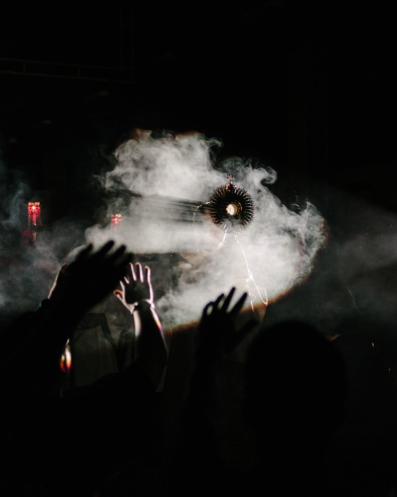 a religious object held up in front of a fog machine with a crowd raising their hands, _fog machine in church 4k