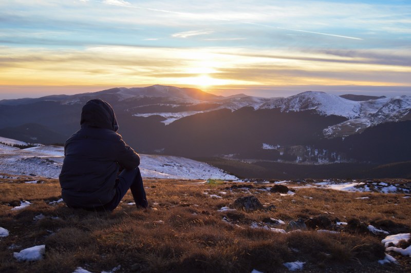 a person sitting atop a mountain in sinaia watching the sunset above snowy mountain range, sunset watching on sinaia mountain 4k
