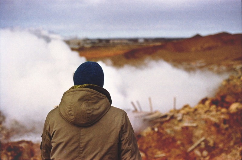 a person in a jacket faces away toward white smoke outdoors in iceland, facing white smoke 4k