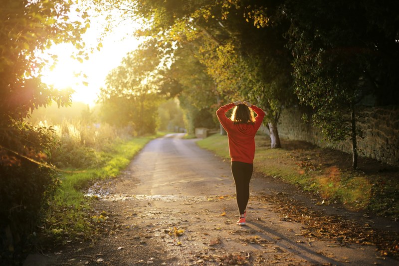 a woman wearing a red sweater and sneakers walking with her hands on top of her head down a path during fall in west sussex, morning jog in the countryside 4k