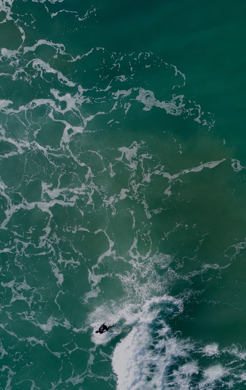 an overhead drone shot of a man in a wetsuit riding a surfboard in the sea at gisborne new zealand, curve 4k
