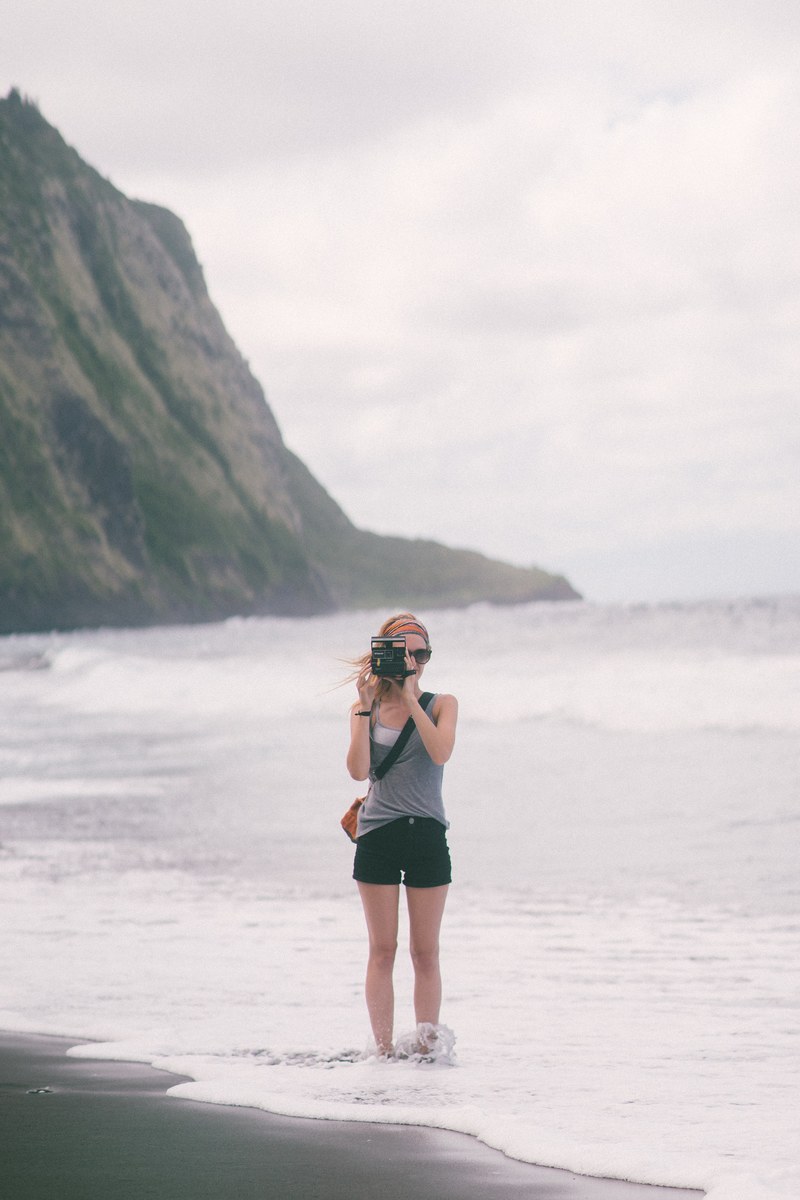 young woman standing on the beach and taking a photo with a polaroid camera, black sands 6 4k