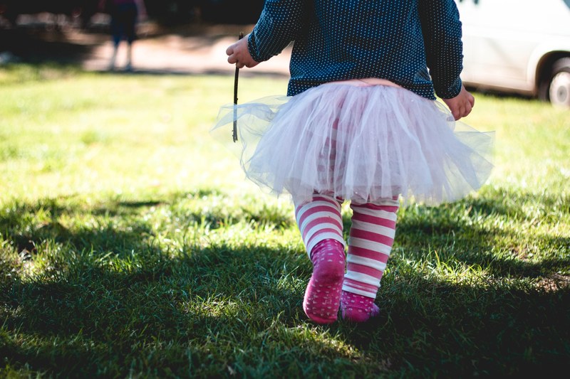 female child wearing a tutu with stripe leggings walks on grass towards sunlight holding a stick, child in tutu_ 4k