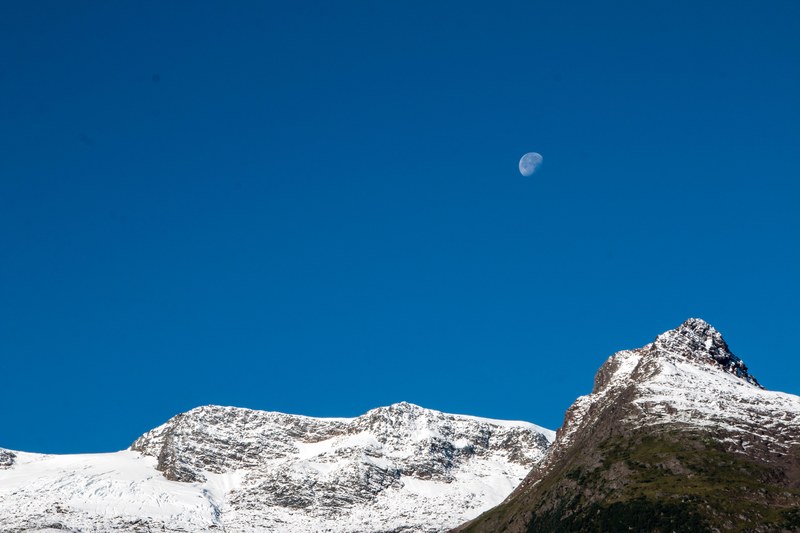 a waning moon in the blue sky above a snow capped mountain ridge, moon above snowy peaks 4k
