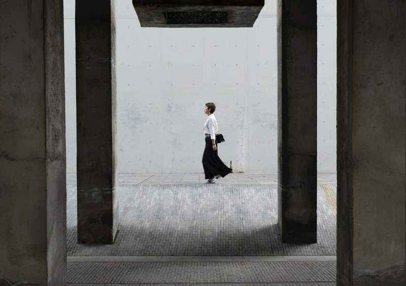 a woman in a black skirt walking on a sidewalk near a wall, architectural frames 4k