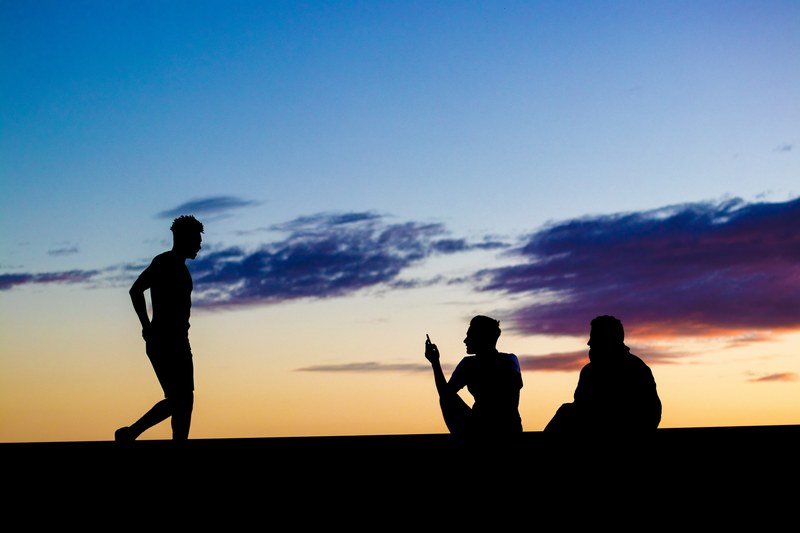 silhouette of a man walking towards two friends sitting under a blue and orange laton sunset, friends 4k