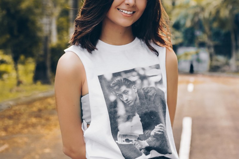 a smiling woman in an arctic monkeys tank top stands on a street by a park, woman in a white tshirt 4k