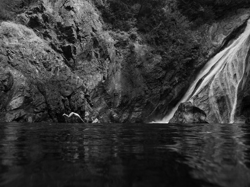 a black and white photo of a person diving into the water under a rock face with a waterfall, diving into the unknown 4k