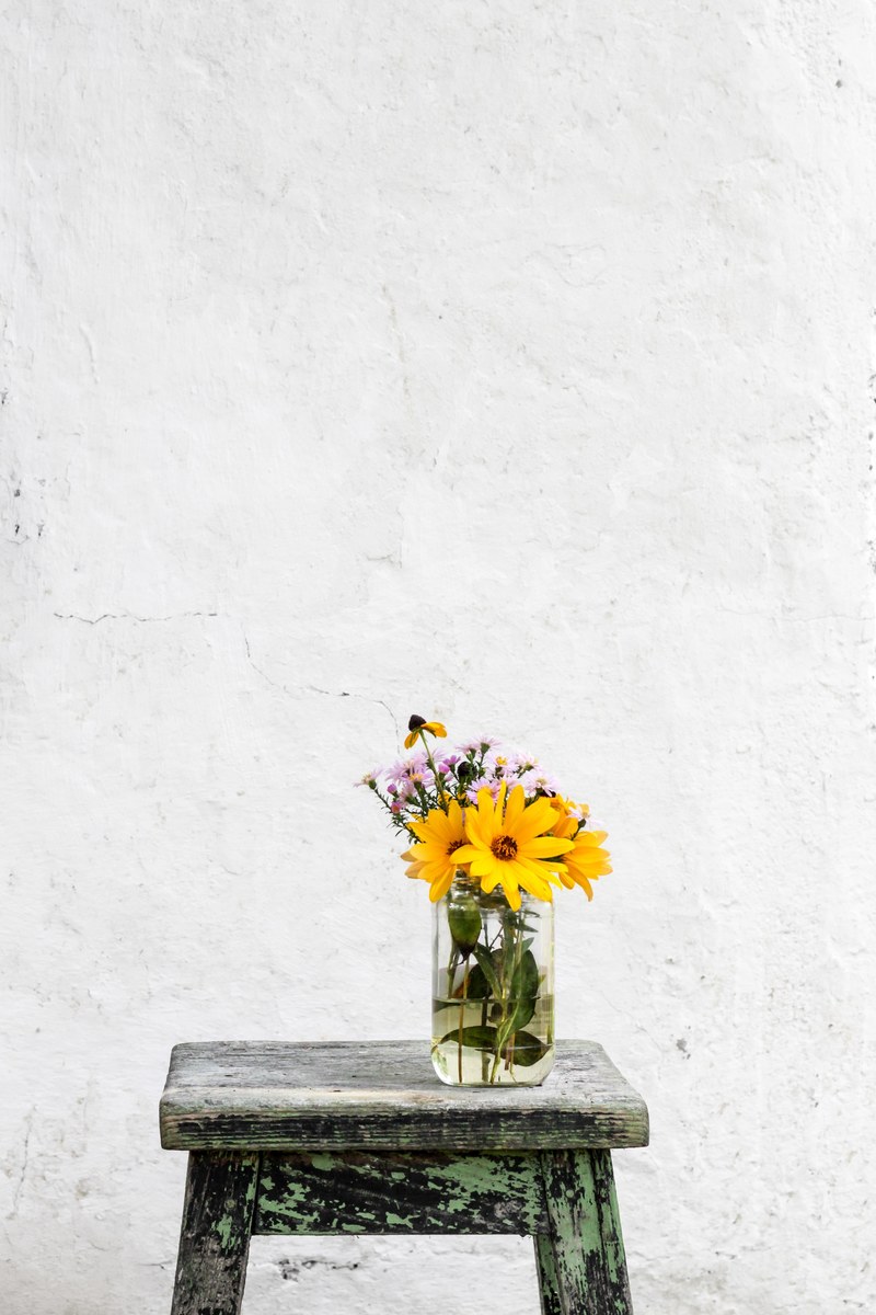 a glass vase with large yellow and small pink flowers on a worn out stool against an old white wall, pink and yellow flower vase 4k