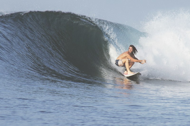 a male surfer with long head wearing board shorts crouching as he rides a cresting wave at sumbawa, crouching surfer in action 4k