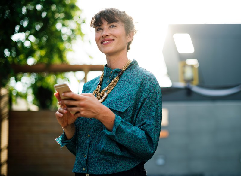 a smiling woman in an azure blouse holding a smartphone while standing outdoors, smiling woman with a phone 4k