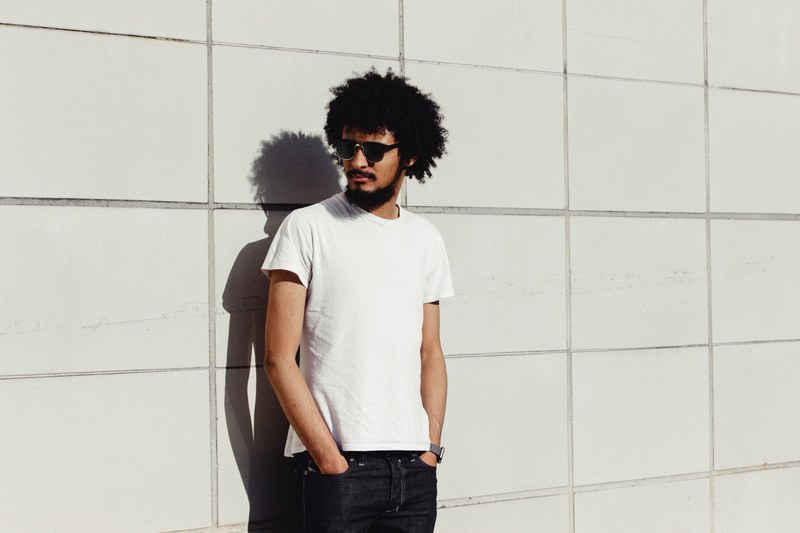 man with curly hair wearing sunglasses standing against a wall, man standing alone 4k