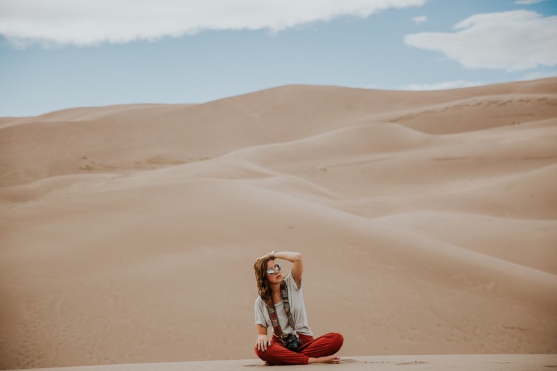 a woman in sunglasses with a camera on a strap sitting cross legged in a desert, sitting in the desert 4k