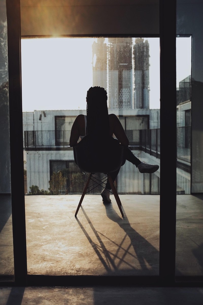 a person facing away from the camera sits in sunshine on a ledge outside a window, sitting outside a window 4k
