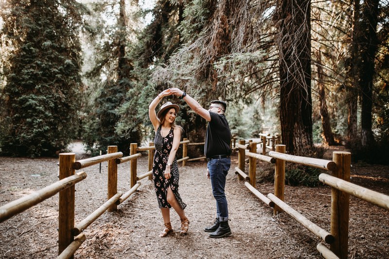 a man and a woman dance holding hands on a fenced in pathway in the woods, surprise proposal in the redwoods 4k
