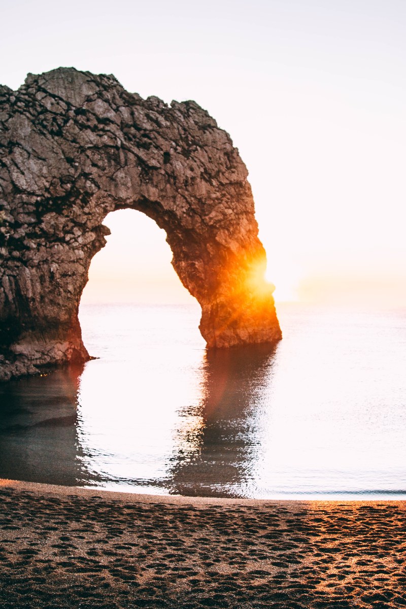 a natural rock arch is illuminated with the rays of a golden sunset over the water near the shoreline of durdle door, dorset sunset sights_ 4k