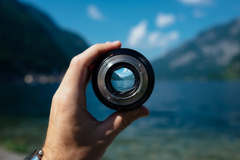 a persons hand holding a camera lens over a mountain lake, mountain lake in camera lens 4k