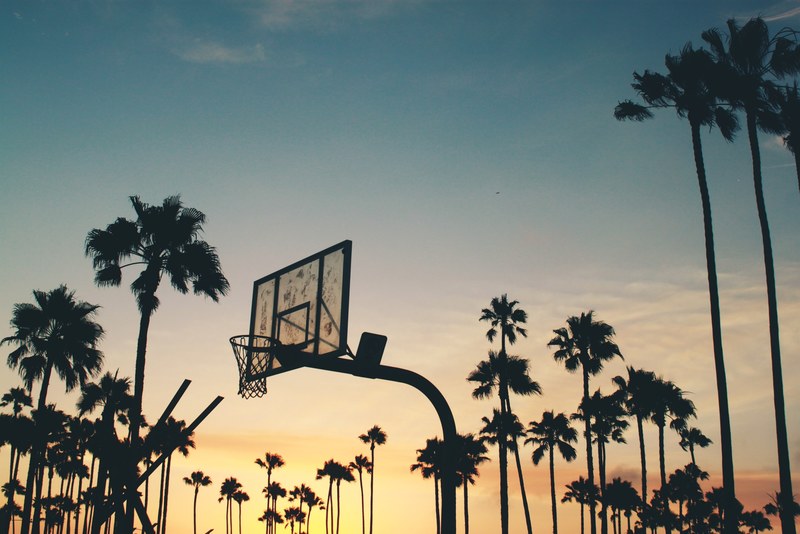 a yellow and blue sunset in venice los angeles forming silhouettes of a basketball court and numerous palm trees, outdoor basketball in venice 4k