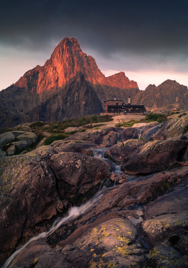 modern house at the top of a rocky hill with mountains and a creek in the background and foreground during sunset, house in the mountain 4k