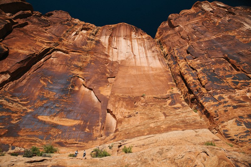two people at the foot of a tall vertical face of red rock, under a tall sandstone face 4k