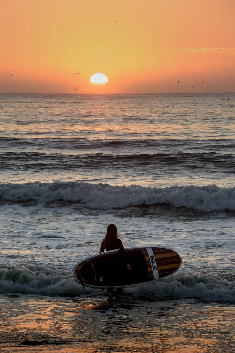 woman surfer holding a surfboard standing in a shallow water during sunset at pacifica state beach, woman surfer during sunset 4k