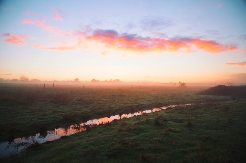 a countryside field with a small stream reflecting the pink blue and orange sky during sunset, countryside sunset 4k