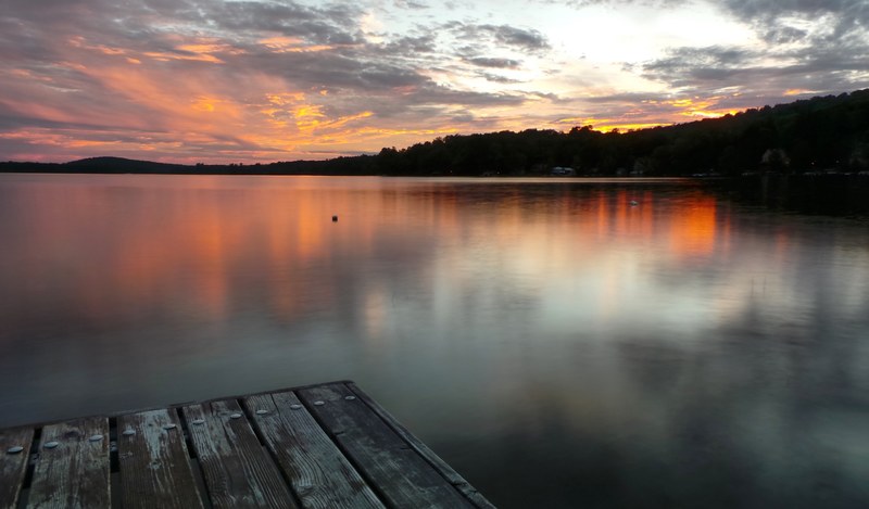an orange and blue sunset reflecting upon a calm lake seen from a wooden pier, calm lakeside sunset 4k