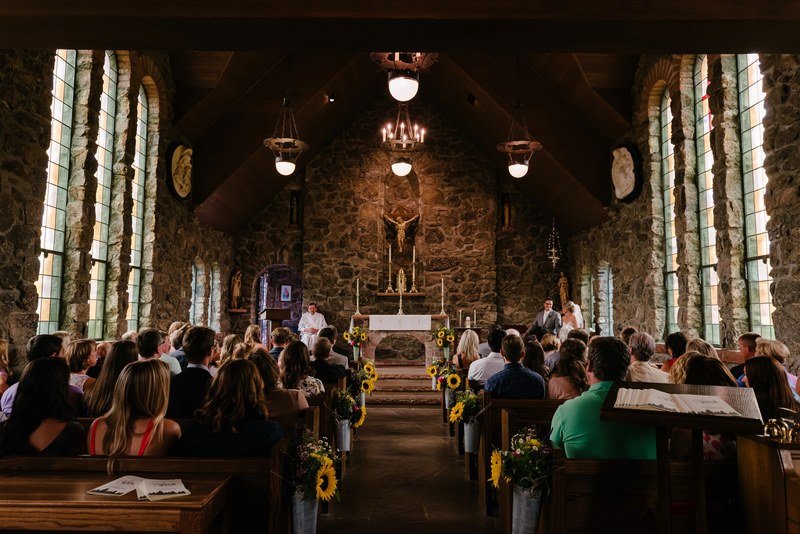 looking up the aisle of a church wedding with guests sitting in the chapel, sunflowers down aisle stone church 4k