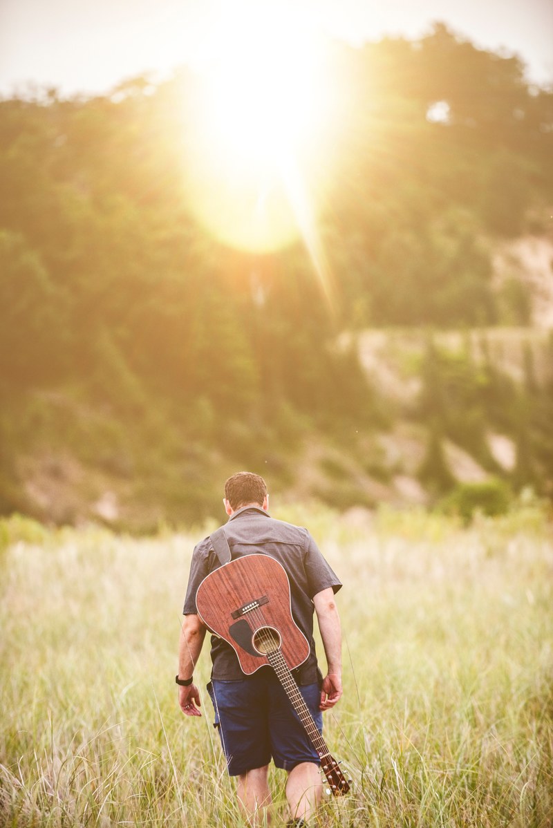 man with acoustic guitar strapped to back walking through a grassy field during sunset, guitar player walking sunset 4k