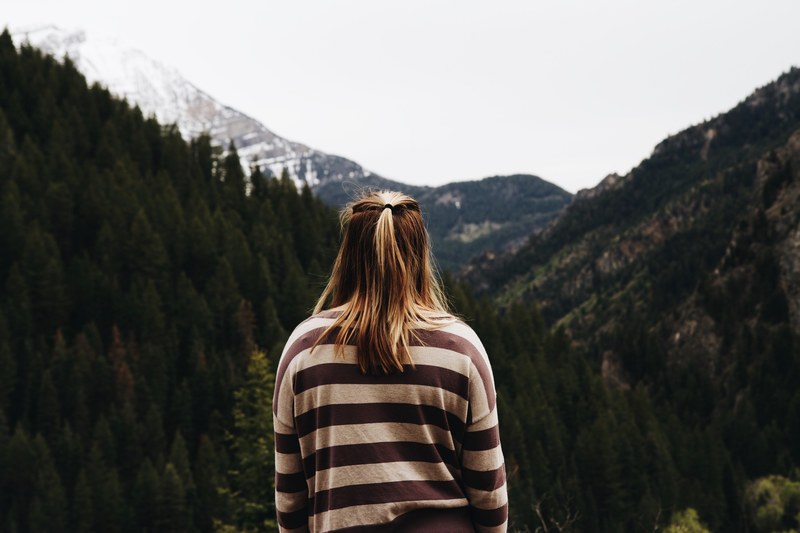 a woman standing back to camera and looking at snowy mountains behind the woods, looking off into the mountains 4k
