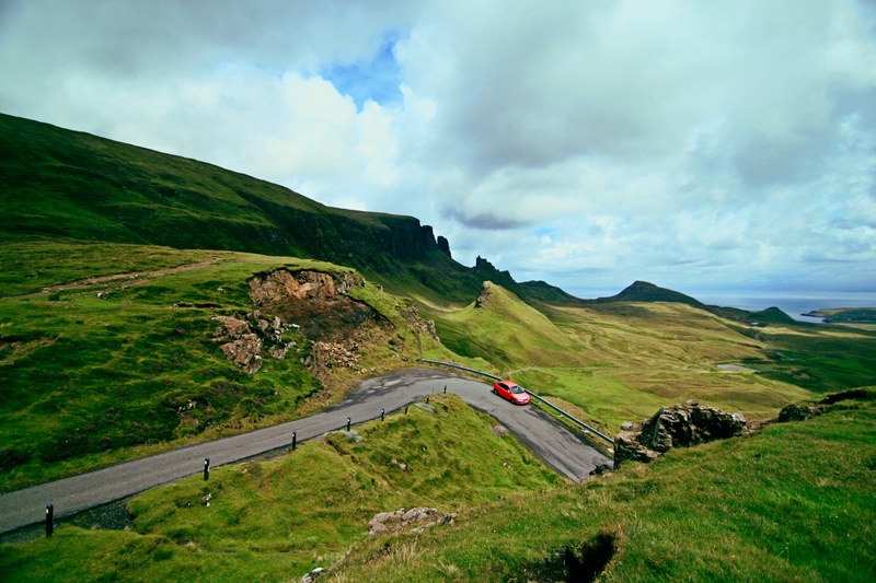 red car driving on countryside road surrounded by green landscape and sloped cliffs, red car countryside road 4k
