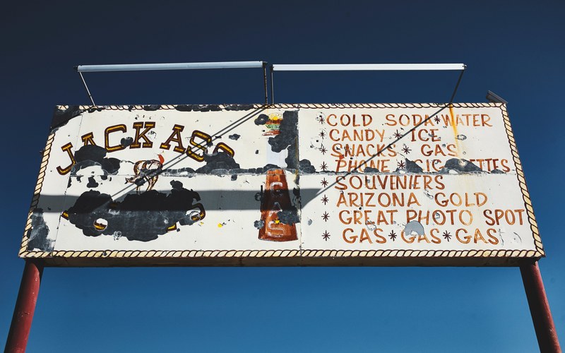 a rusty old signboard about jackass gas station on the road, jackass signboard on road_ 4k