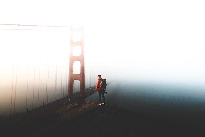 a man with a backpack standing on top of a ledge above a large suspension bridge, _fogust in san francisco 4k