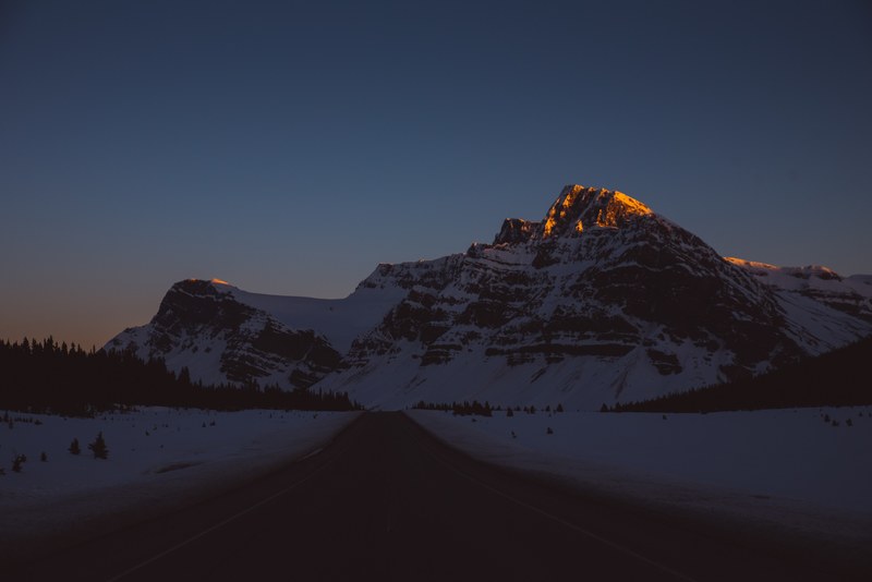 a dim shot of an empty asphalt road running towards snow covered mountains, mountain road in the evening 4k