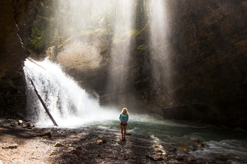 a woman standing near a waterfall tumbling down into a stream, woman near a frothy waterfall 4k