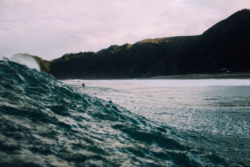 a shot of a rolling wave with a surfer in the distance and the mountains in the background at piha, surfing in the shadow of the mountains 4k