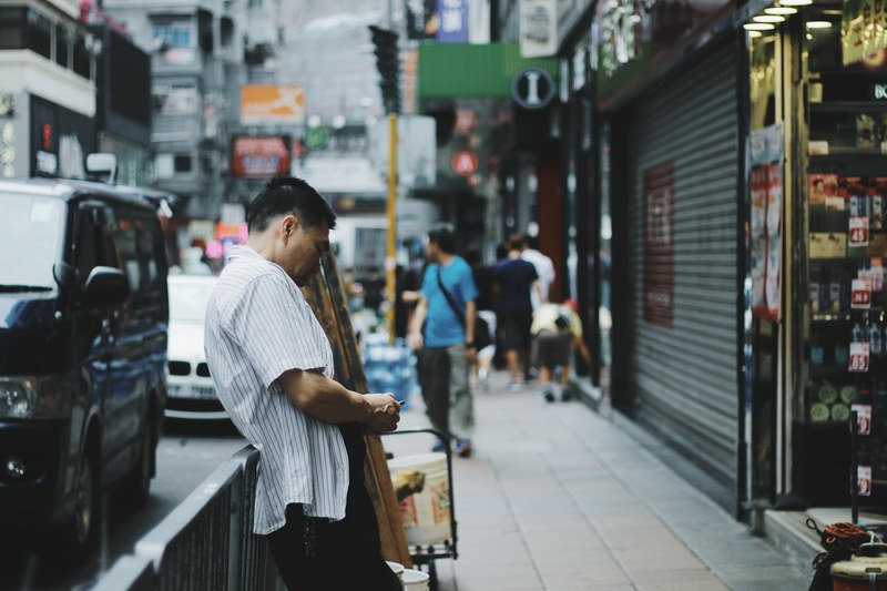 a man on the sidewalk looks down aside a city street and shops in hong kong, man aside city street 4k